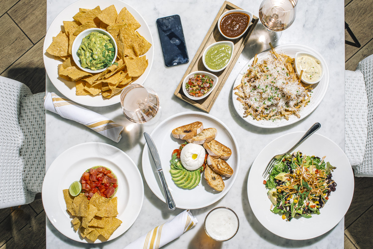 overhead photo of a table with a variety of food including burrata, chips and guacamole and truffle fries