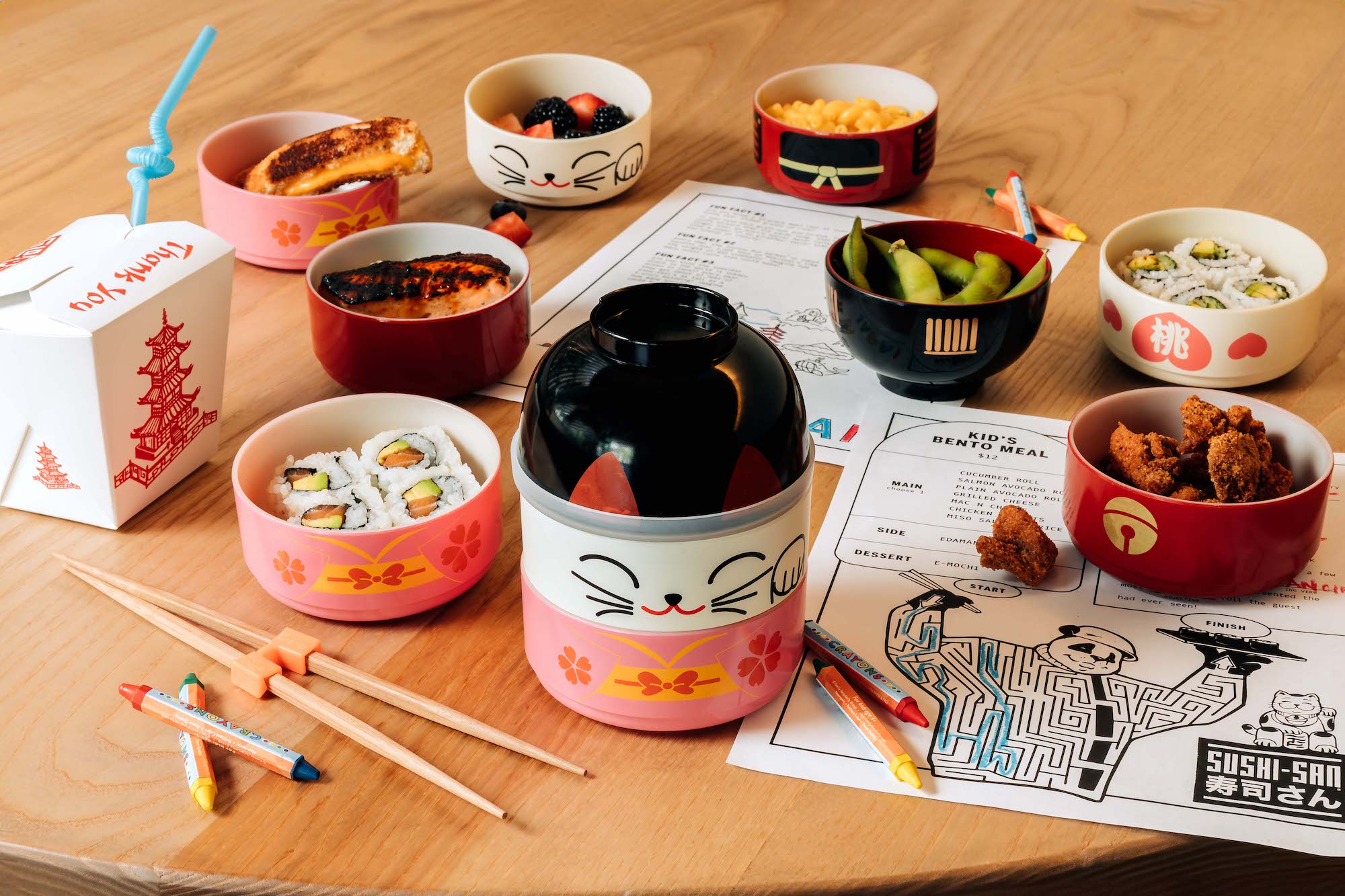 From left to right: a takeaway rice box, children’s chopsticks, salmon avocado roll, cooked salmon, a kitten bowl, edamame, chicken nuggets and a cucumber roll with kids menus on the table