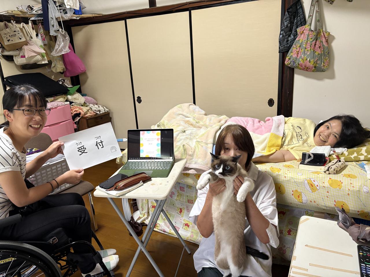CIL松江代表中村さんの自宅リビングにて、ピアカウンセリングの準備をしているところ。中村さん、「受付」の紙をもった曽田、介助者とネコさんが笑顔で。