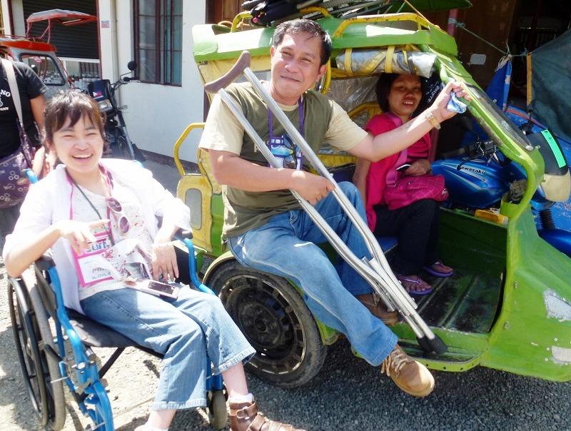 フィリピンの村役場の前で、左から笑顔のMさん、松葉杖をついた現地サポーターの男性、フィリピンの三輪バイクタクシーに乗っている現地の障害女性がみんな笑顔で。