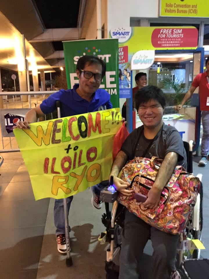 イロイロ空港で、「welcome!」という手書きの旗を掲げる松葉杖のスタッフと、笑顔の男の子
