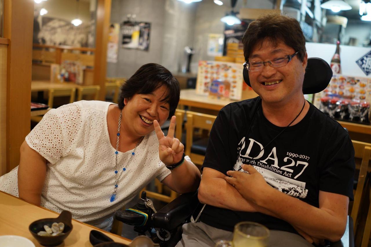 居酒屋にて、笑顔でピースする盛上さんと、横で笑う車いすユーザーの今村さん