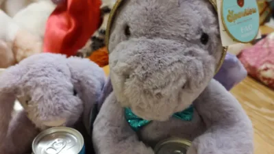 A purple bunny with starry ears is sitting next to a purple dragon wearing a sparkly gold and blue party hat and a sparkly blue bowtie. They are both holding cans of WKD. King Purple Bunny is behind them, out of focus.