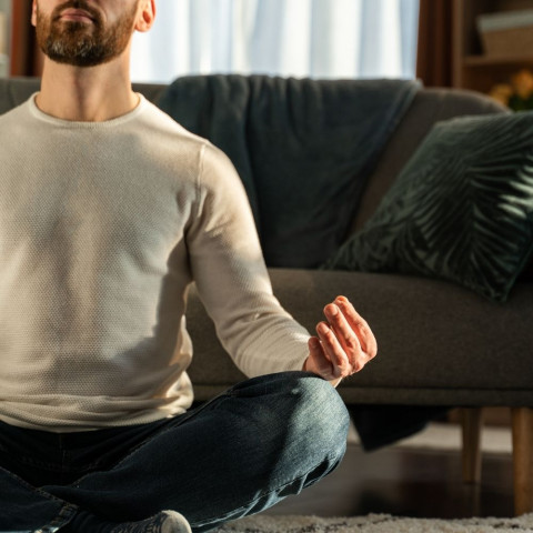 Muškarac ispred kauča meditira u trapericama i džemperu