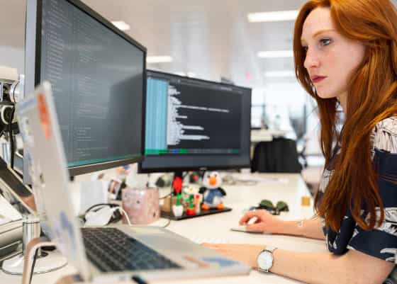Uma mulher ruiva em frente a duas telas de computador. Em uma das telas, um terminal de computador está aberto.