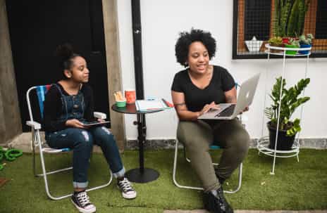 Duas pessoas estão em um jardim com uma grama verde. Do lado esquerdo, uma adolescente com um macacão jeans, tênis branco e branco, blusa preta está sentada em uma cadeira colorida. Ao lado dela, uma mesa pequena preta com um caderno em cima e copos. Do lado direito, uma mulher com uma blusa preta com manga, calça verde escura está com um notebook no colo, olhando fixamente para ele e sorrindo. Ao fundo, plantas em vasos coloridos.