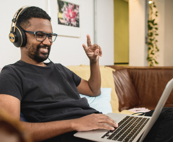 Um homem vestido com a camisa preta, calça preta, óculos escuros com uma armação preta e um fone no ouvido está sentado em um sofá marrom. Ele está com um notebook em seu colo, uma das suas mãos no notebook e a outra fazendo o símbolo de paz e amor com os dedos. No fundo, uma parede branca com quadros pendurados na parede.