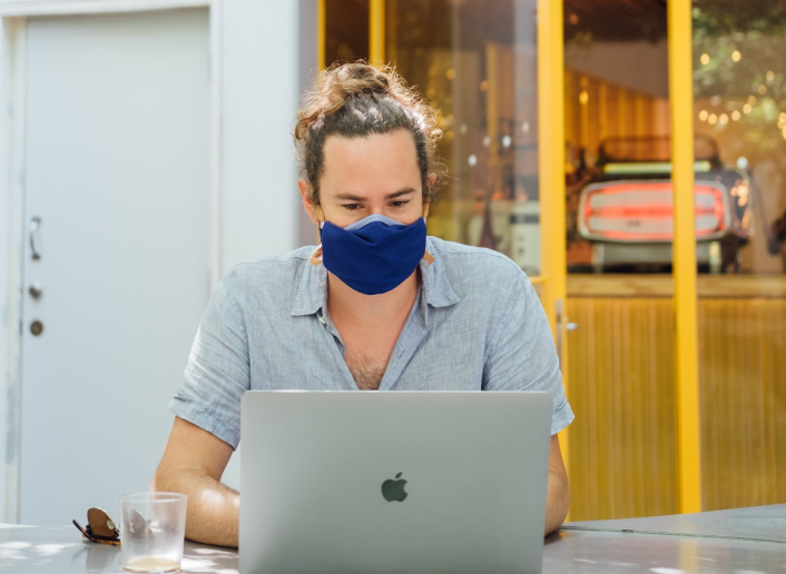 Um homem sentado em uma mesa está olhando para um notebook a sua frente, do seu lado direito tem um óculos de sol na cor marrom e um copo de vidro transparente. O rapaz é de cor clara, cabelo longo mas está preso, usa máscara na cor azul, usa uma camisa de botão na cor azul claro e está com alguns botões abertos. Atrás dele possui uma porta branca fechada, e do lado uma porta de vidro na cor amarela.