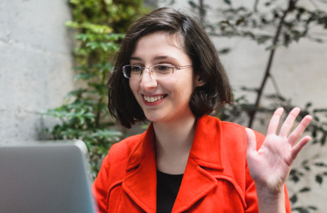 Na imagem uma mulher que está usando uma blusa vermelha está olhando para o notebook enquanto levanta a mão. Ela está rindo. Ao fundo está uma planta.