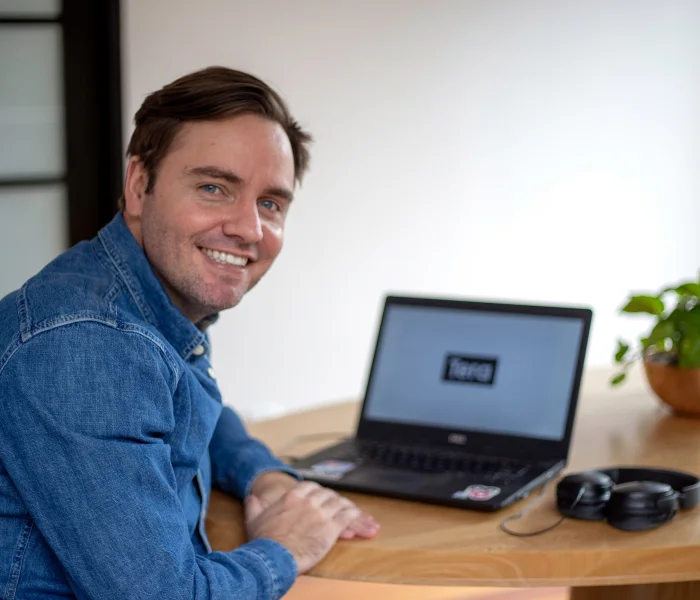 Um homem de óculos segura um notebook preto, enquanto mexe no trackpad. Ele usa uma camiseta verde musgo, um óculos quadrado e prata. Tem barba. Ao fundo, desfocado, um homem observa uma parede com vários postits, rosas e laranjas, colados.