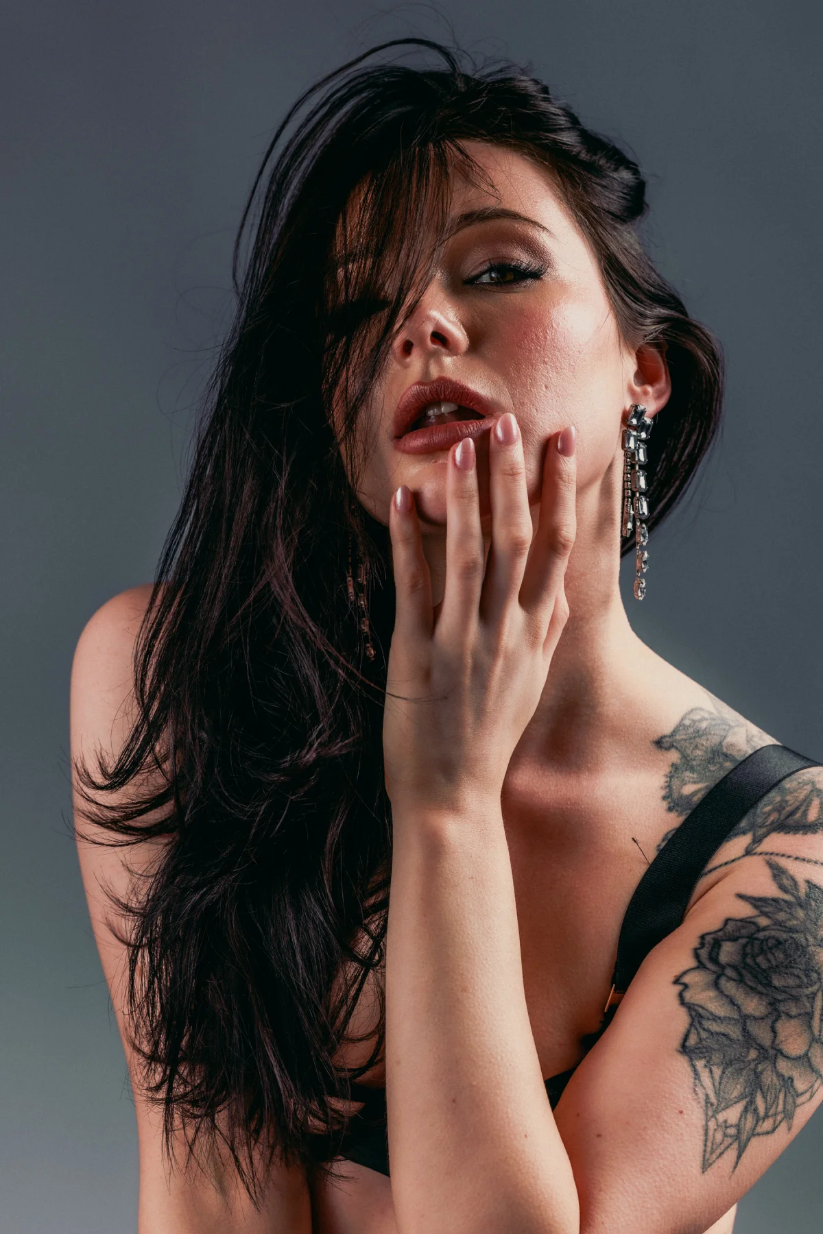 Close-up portrait of a woman with dark hair and a shoulder tattoo, posing against a grey background.