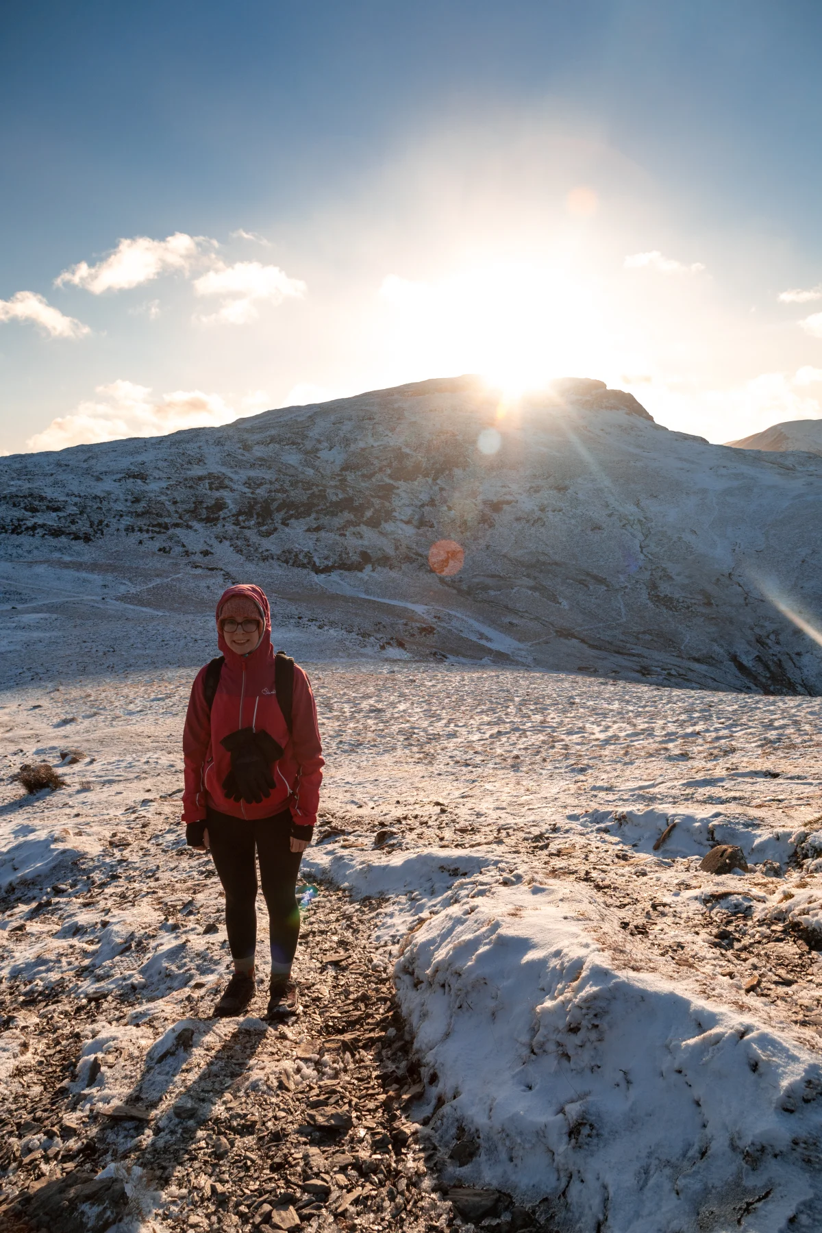A woman in a red jacket standing in a snowy, sunlit mountain landscape.