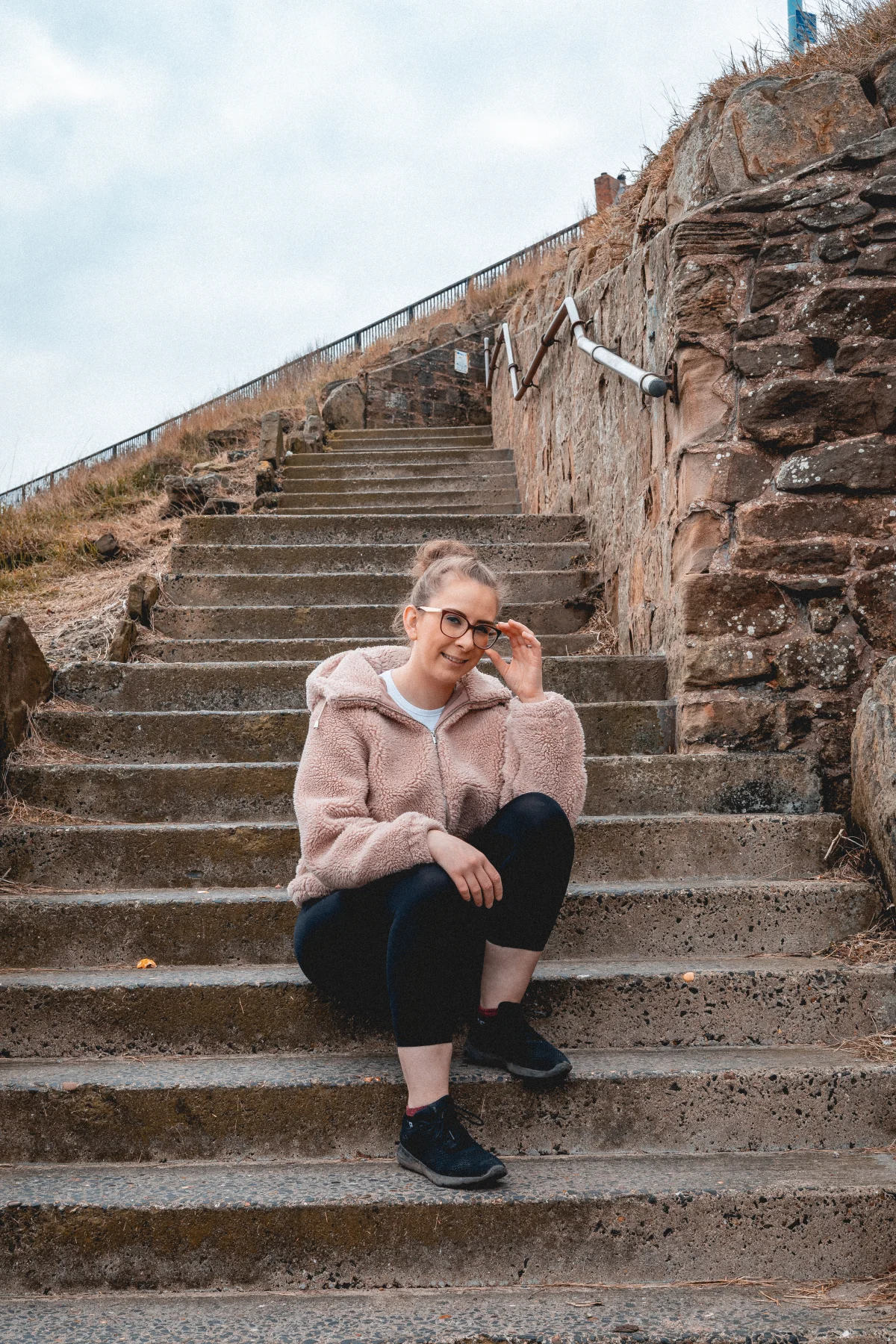 A woman in a pink fuzzy jacket and black leggings sitting on stone steps outdoors.