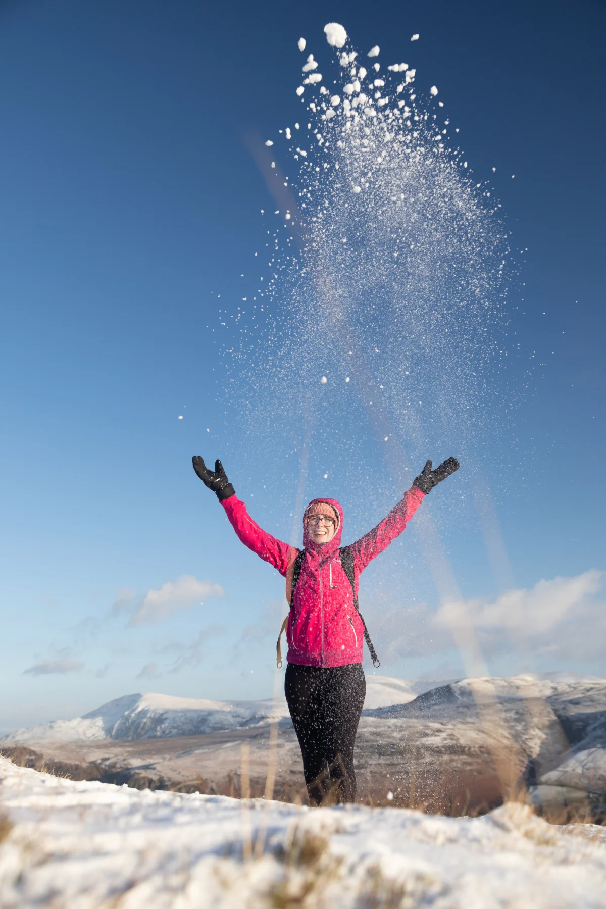 A person in a pink jacket joyfully tosses snow into the air amidst snowy mountains.
