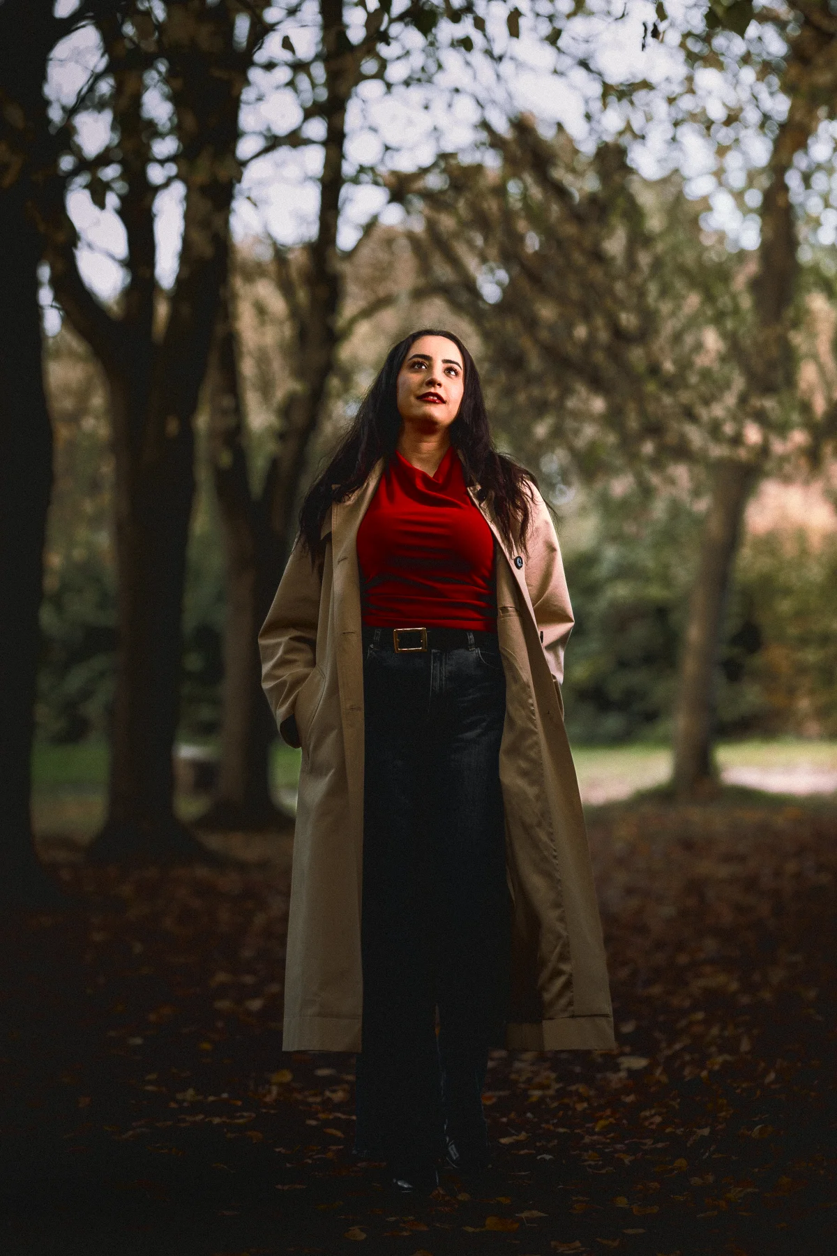 Woman in a red top and beige trench coat standing in a park with fallen autumn leaves.