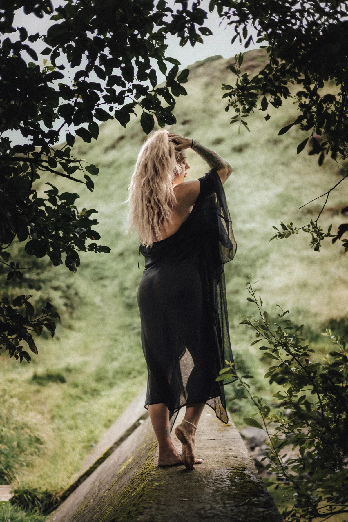 Woman in a sheer black dress standing on a stone ledge, framed by green leaves against a grassy hill.