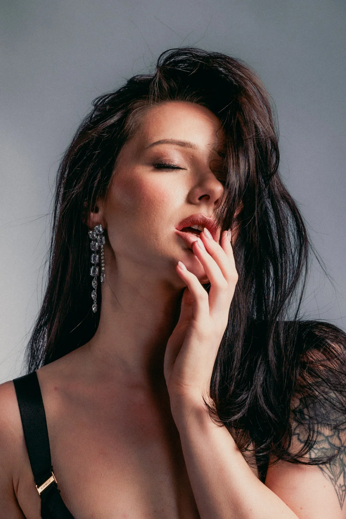Close-up portrait of a woman with dark hair and large earrings, eyes closed, hand near her lips.