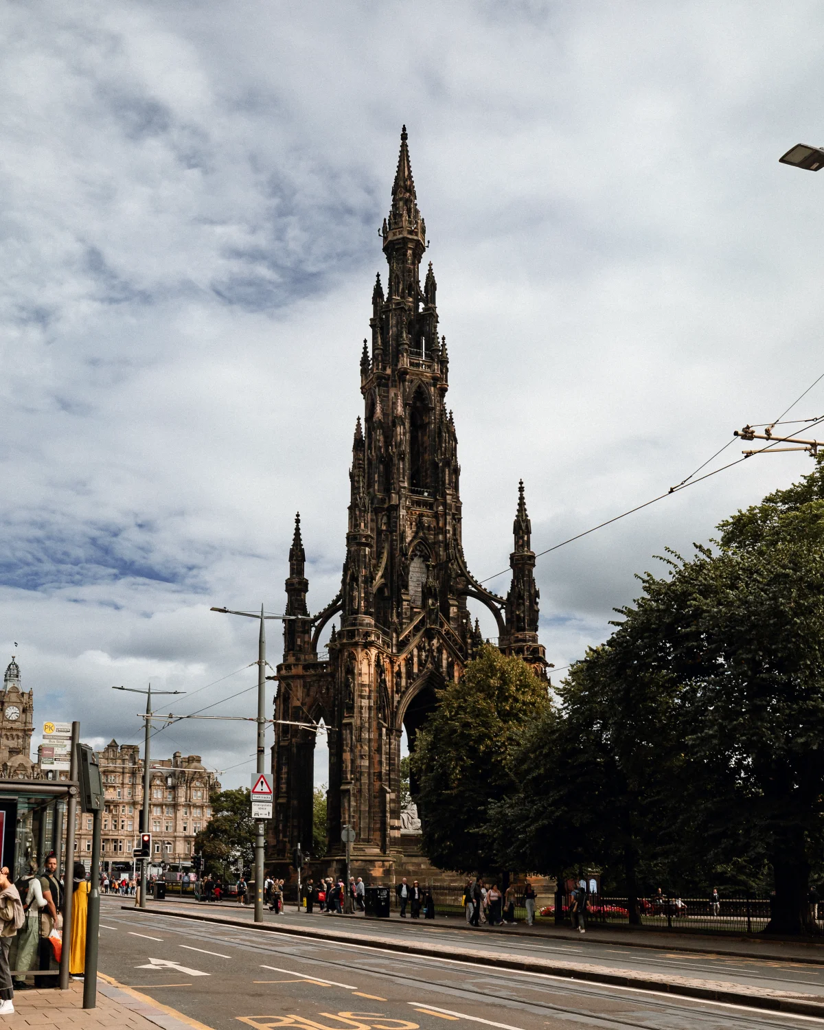 A tall Gothic monument towers over a city street under a cloudy sky, surrounded by trees and urban architecture.