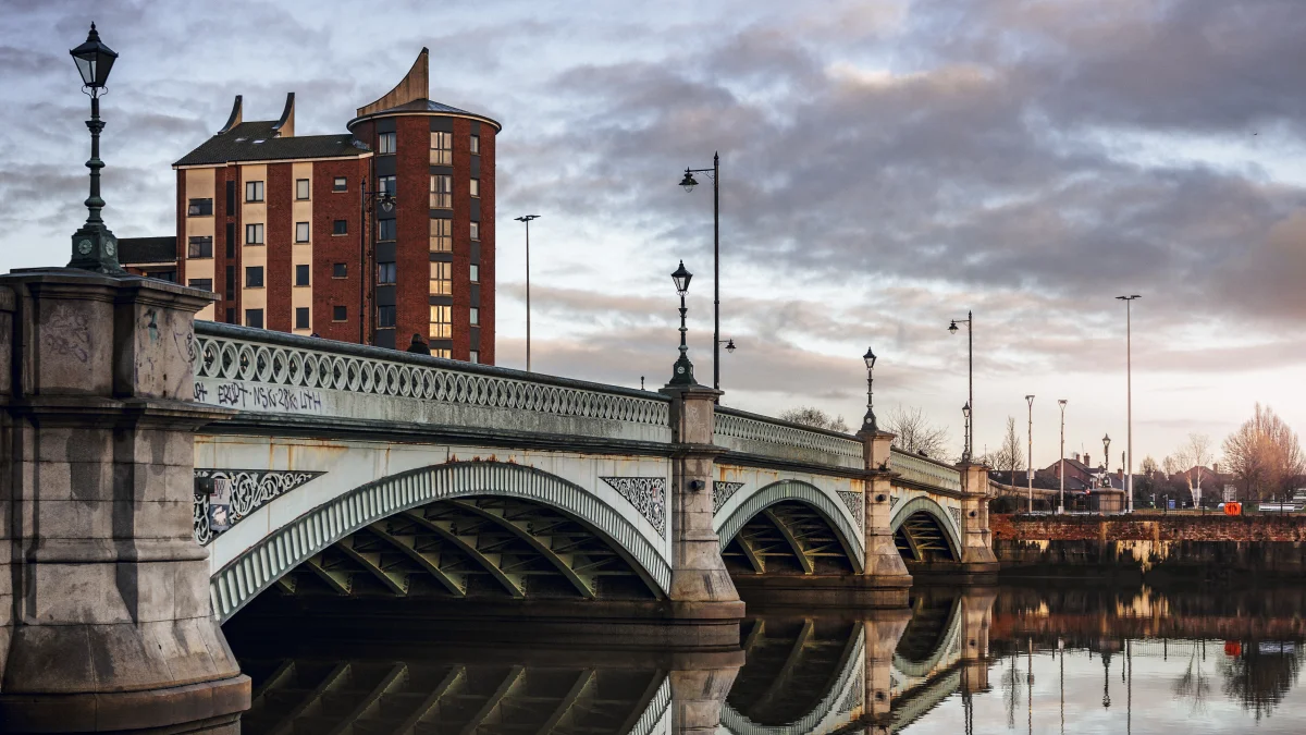 An arched stone bridge over a river with a tall brick building and cloudy sky in the background.