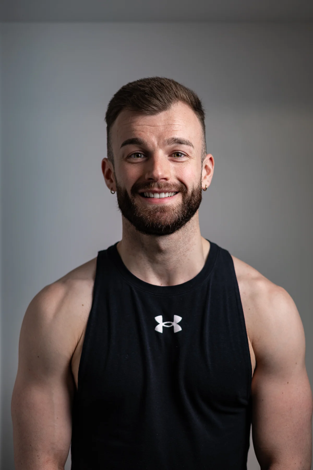 Close-up portrait of a smiling man with a beard wearing a black athletic tank top against a grey background.