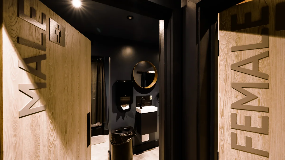 View through open wooden doors into a dark, modern restroom with male and female signage.