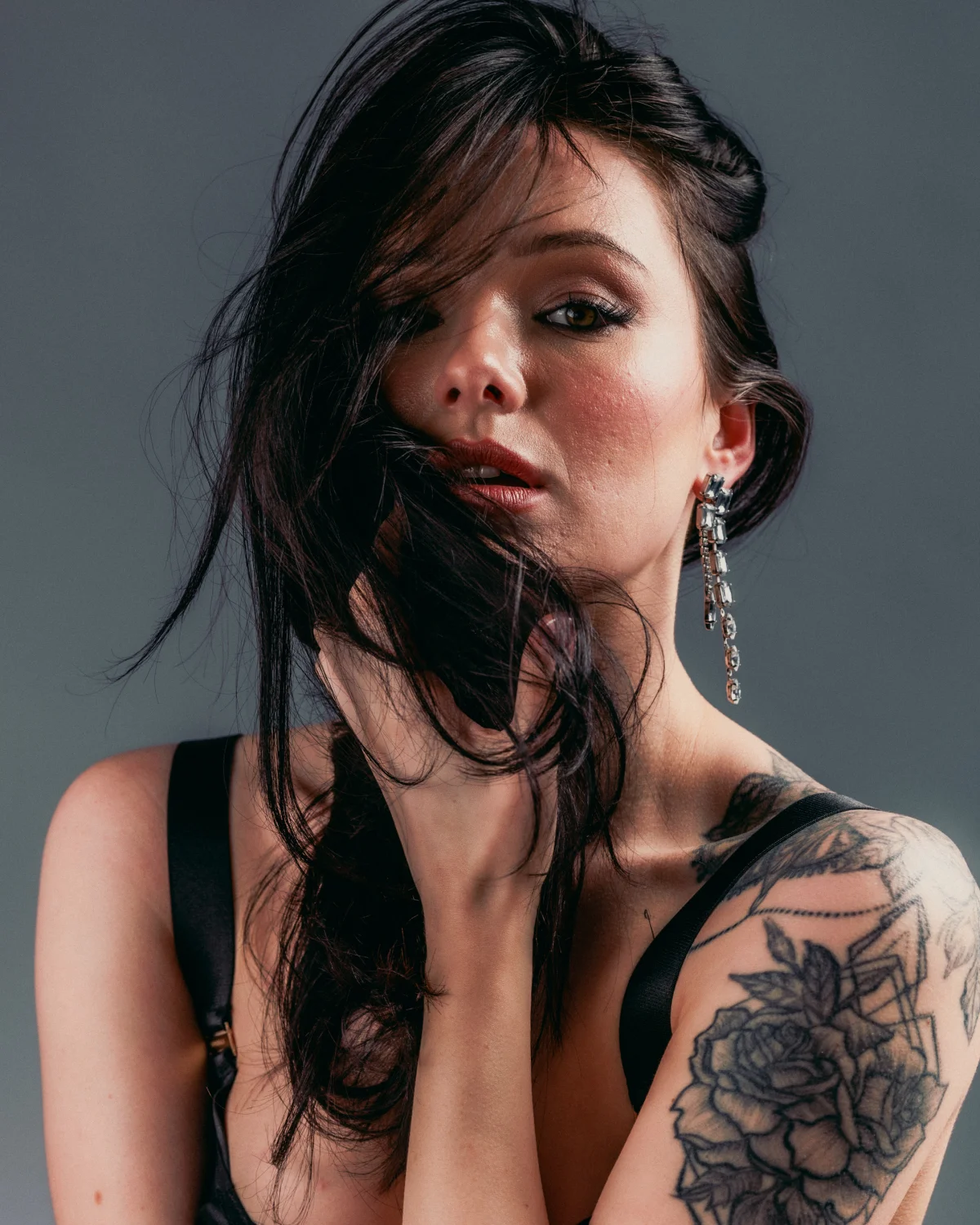 Close-up portrait of a woman with dark hair partially covering her face and shoulder tattoos against a grey background.