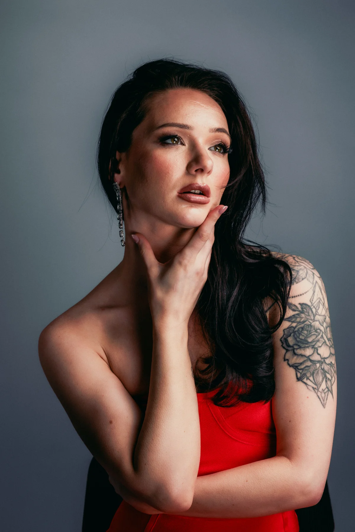 Close-up portrait of a woman with dark hair and a shoulder tattoo, wearing a red top against a gray background.