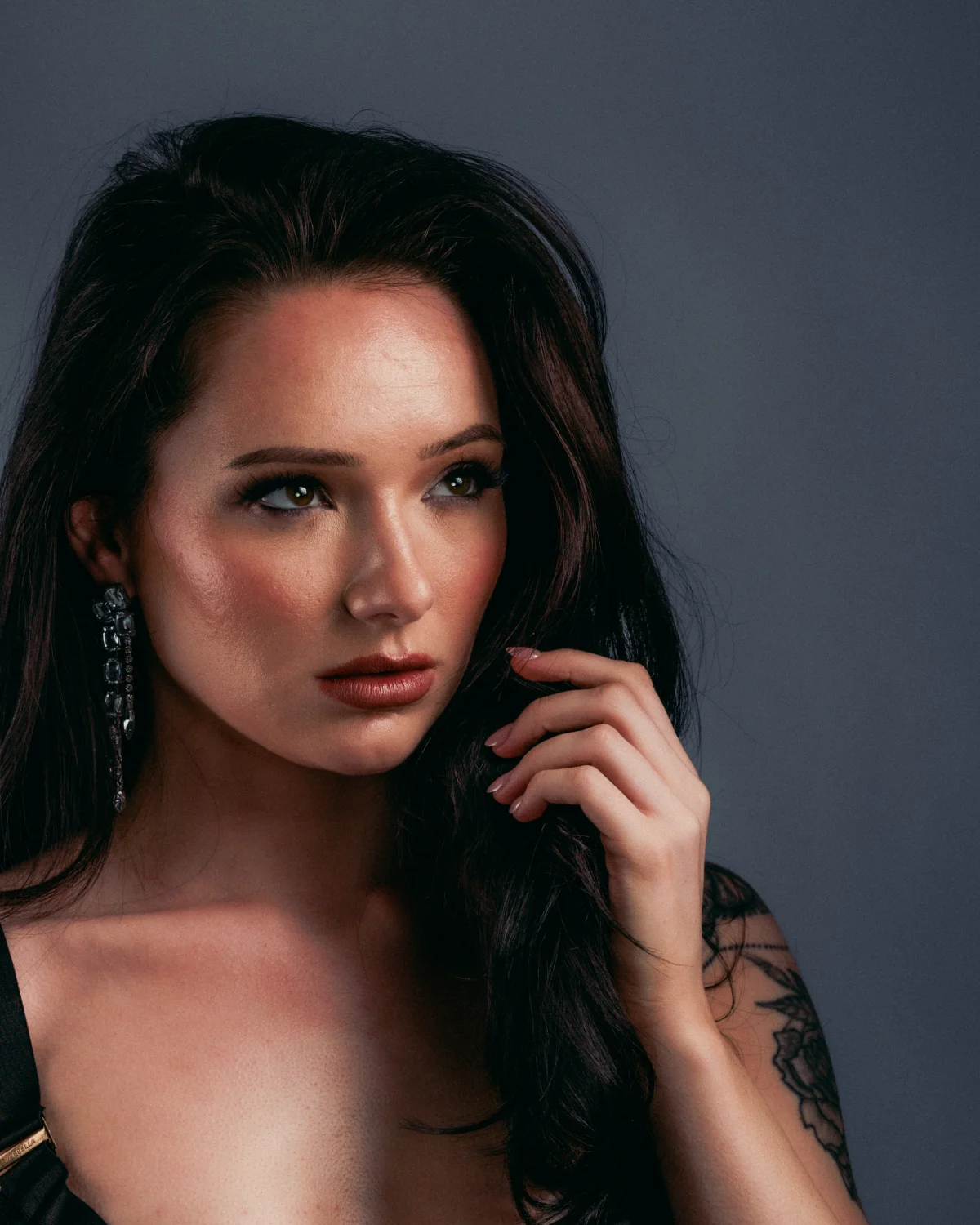 Close-up portrait of a woman with dark hair and a tattoo against a grey background.