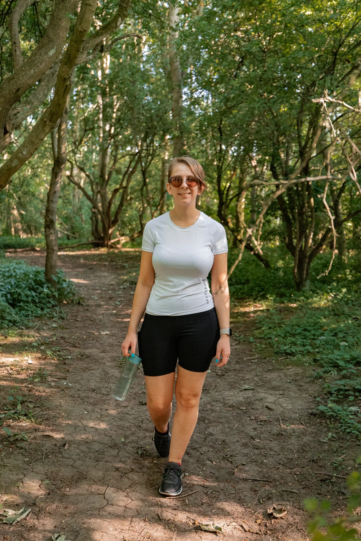 A woman in athletic wear walking along a sunlit forest path, holding a water bottle.