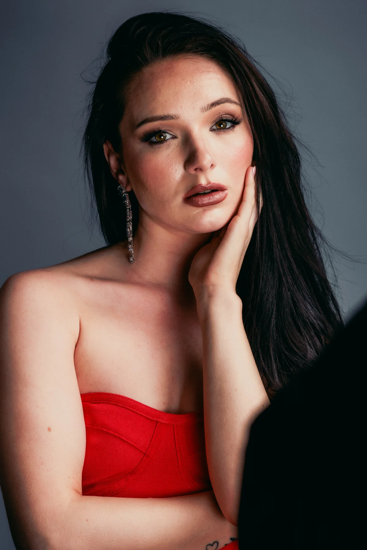 Close-up portrait of a woman with dark hair and a red strapless top against a gray studio background.