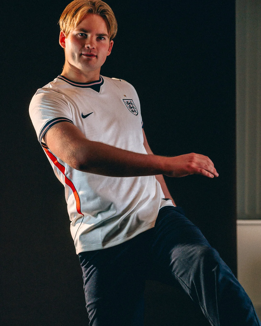 A young man in an England football jersey and navy trousers posing against a dark background.