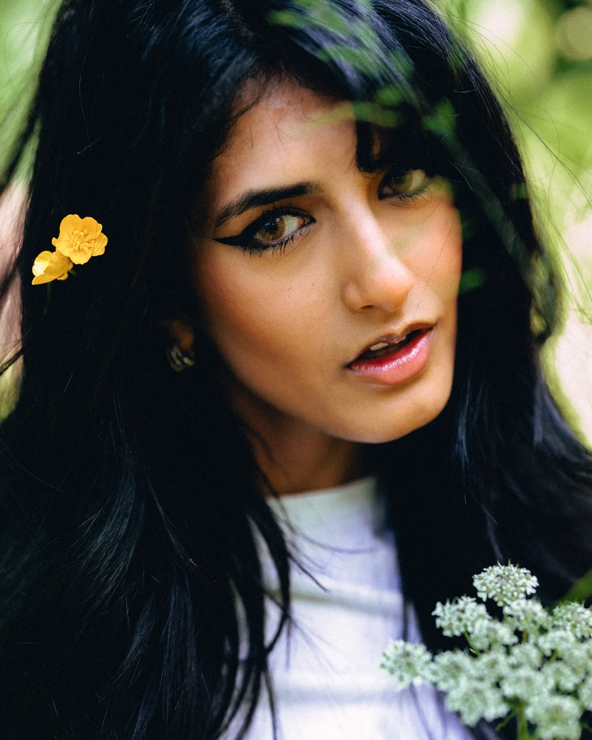 Close-up portrait of a woman with dark hair and winged eyeliner, featuring a small yellow flower in her hair.