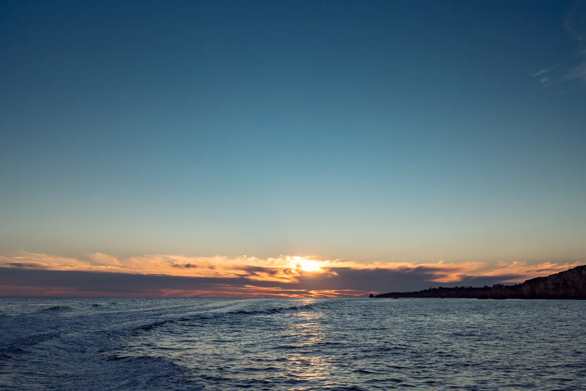 A serene sunset over a calm ocean with golden light breaking through clouds and distant cliffs.