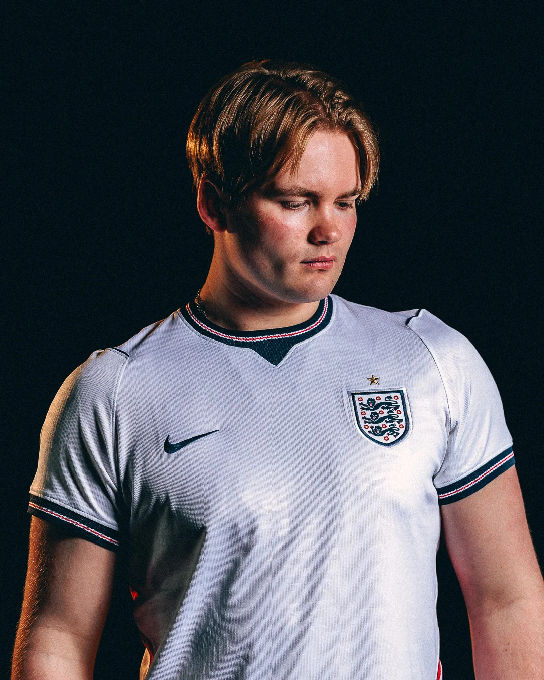 Moody studio portrait of a young man in an England football jersey looking downwards.