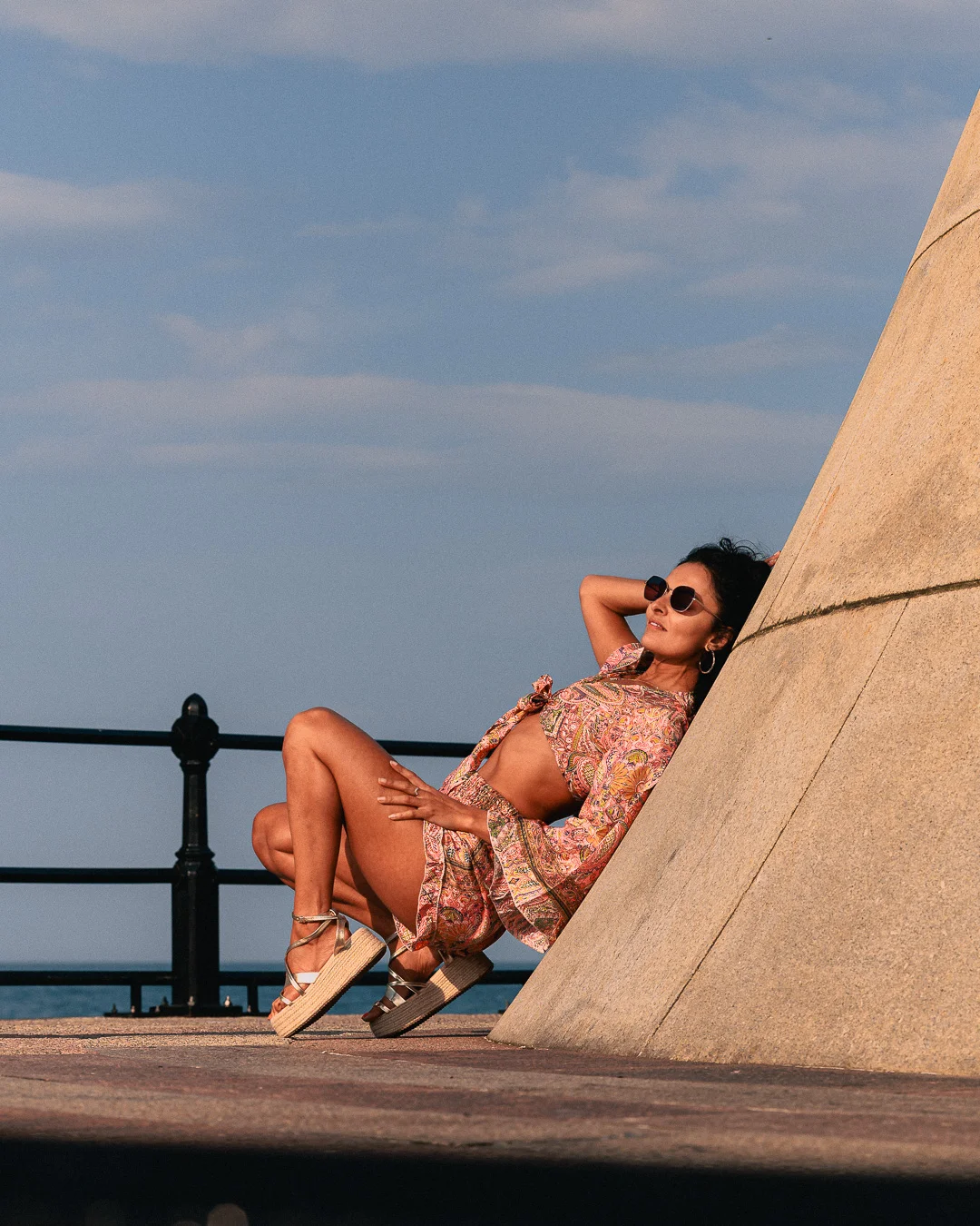 A woman in a colorful patterned dress and sunglasses reclining against a concrete wall by the sea.
