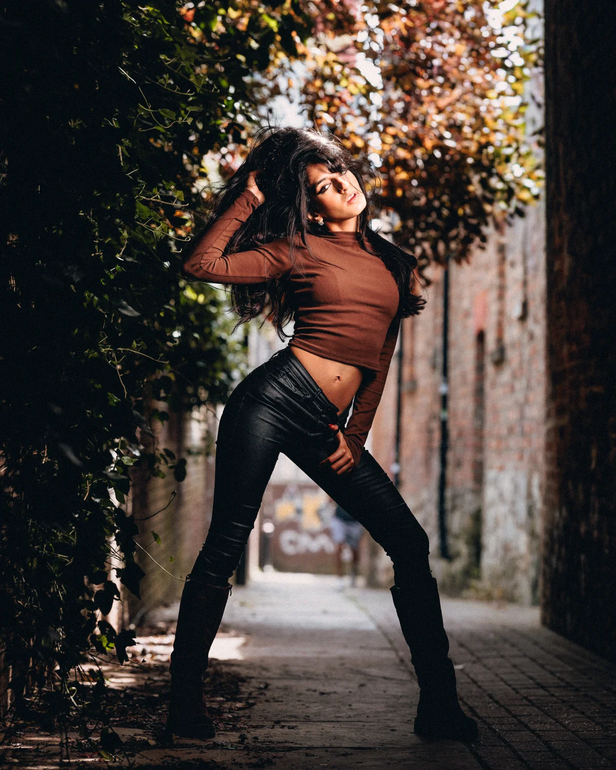 A woman with long dark hair posing in a brown crop top and black leather pants in an urban alleyway with greenery.