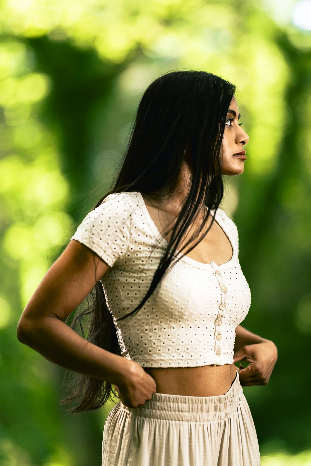 A woman with long black hair in a white cropped top and skirt posing in profile against a blurred green forest.