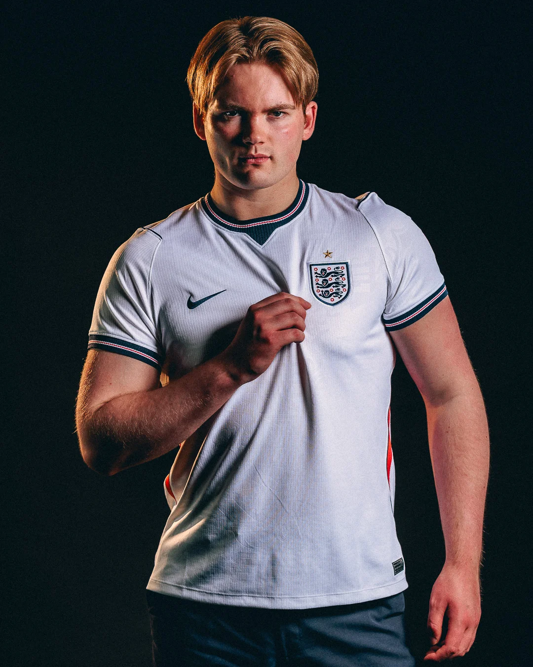A young man with blond hair wearing an England national football team jersey against a dark studio background.