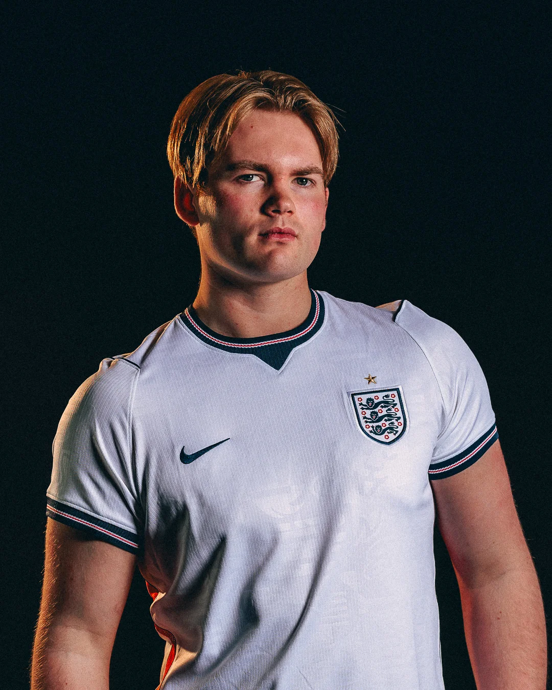 Studio portrait of a blonde man wearing an England national football team jersey against a black background.