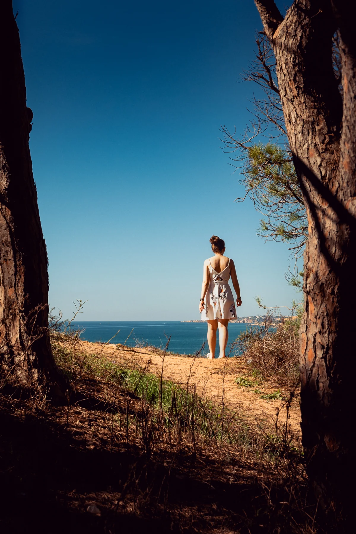 Woman in a white dress standing on a cliff overlooking the blue ocean, framed by two large tree trunks.