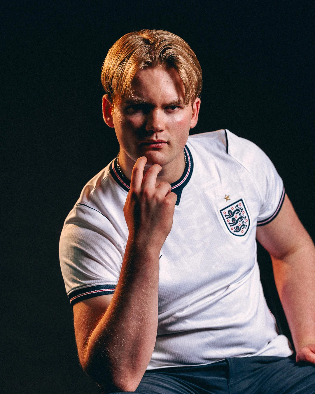 A young man with blond hair in a white England football jersey posing thoughtfully against a dark background.