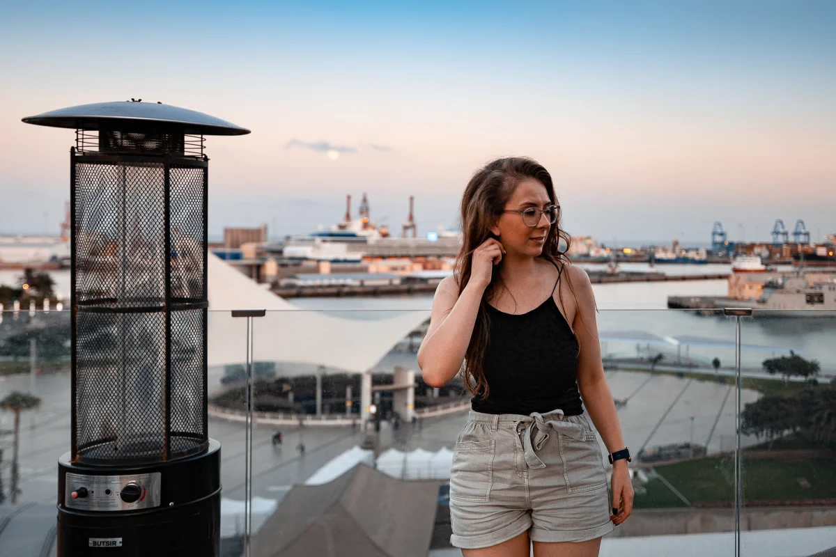 Woman in black tank top and beige shorts posing on a balcony with a port city background at dusk.