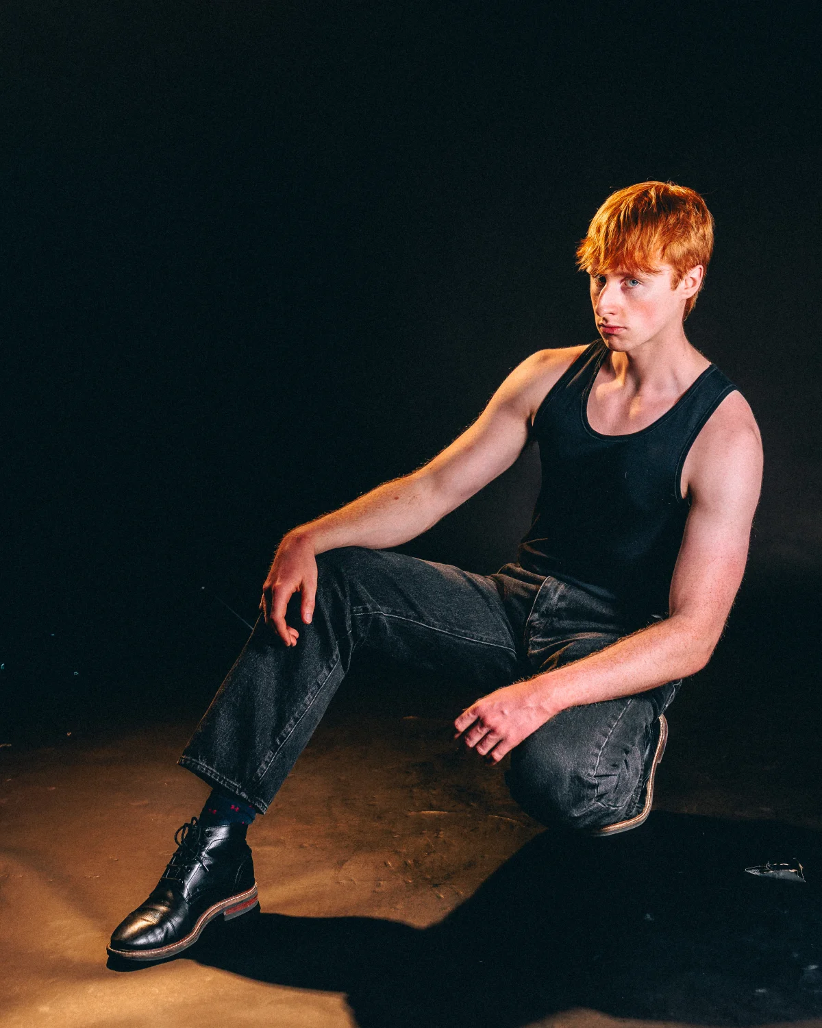 A red-haired man in a black tank top and dark jeans sitting on a dark floor against a black background.