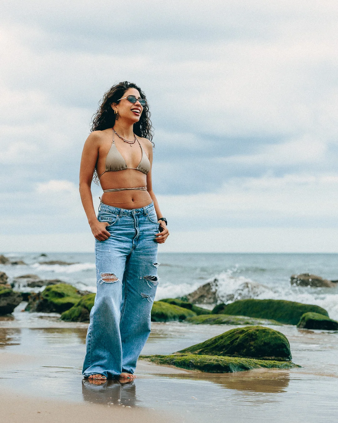 A smiling woman in a gold bikini top and ripped blue jeans posing on a beach with mossy rocks.