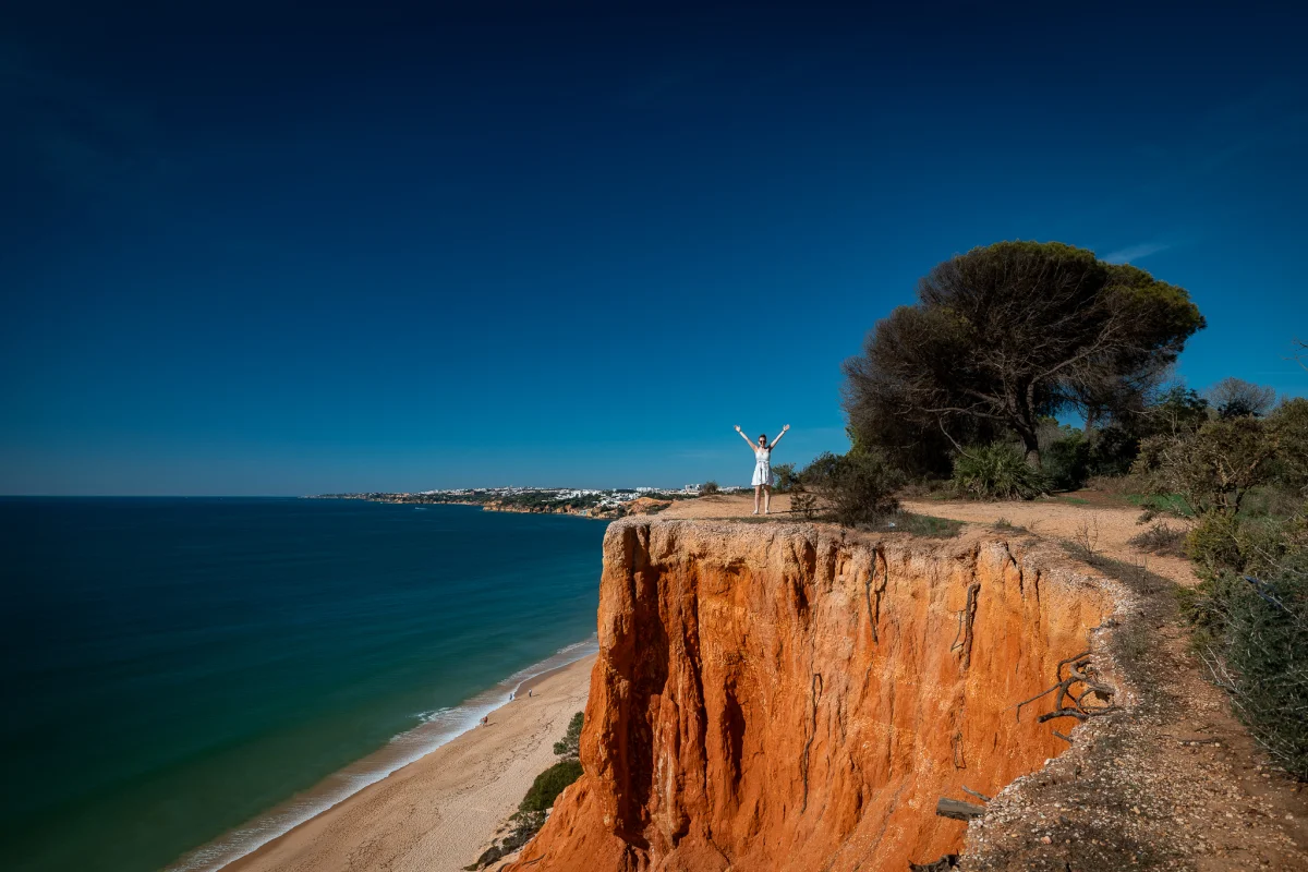 A woman in a white dress stands with arms raised on a red cliff overlooking a blue ocean and sandy beach.