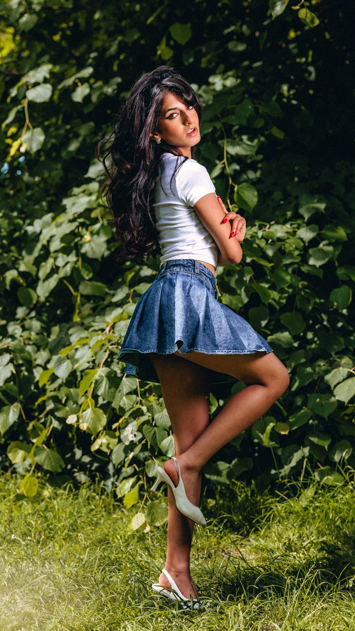 Woman with long dark hair posing in a white t-shirt and short denim skirt against lush green foliage.