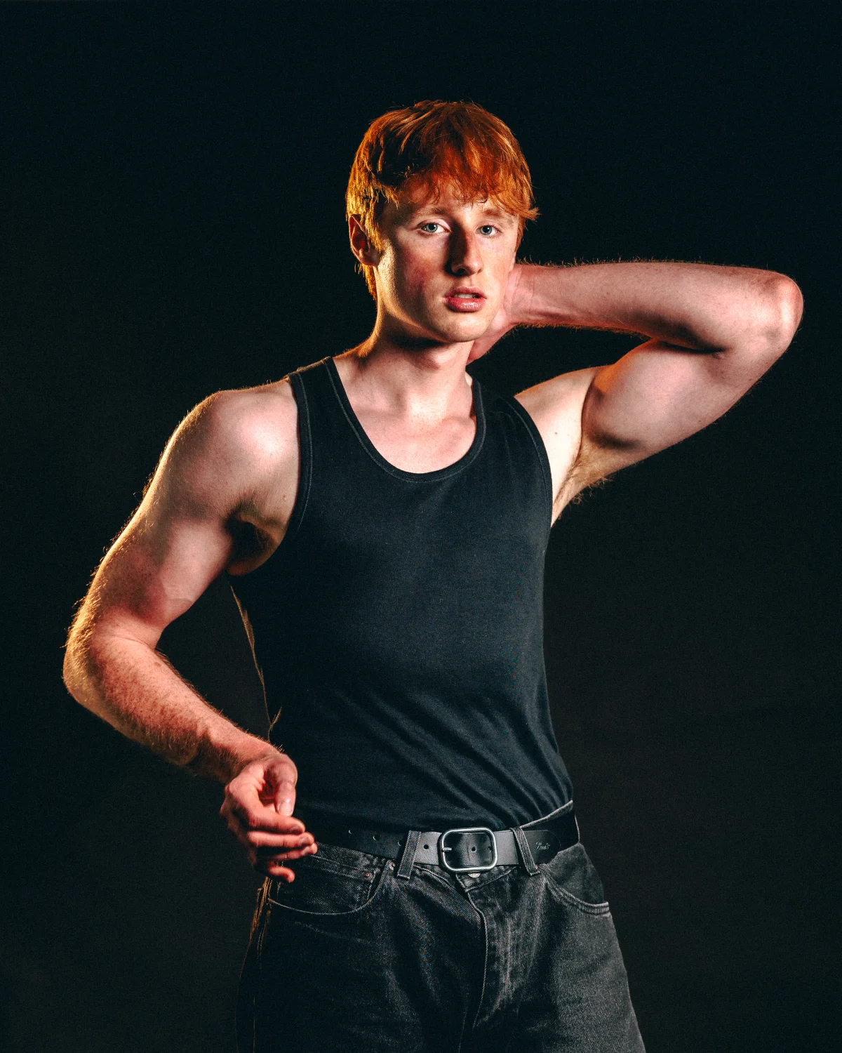 A red-haired man in a black tank top and dark jeans posing against a solid black background.
