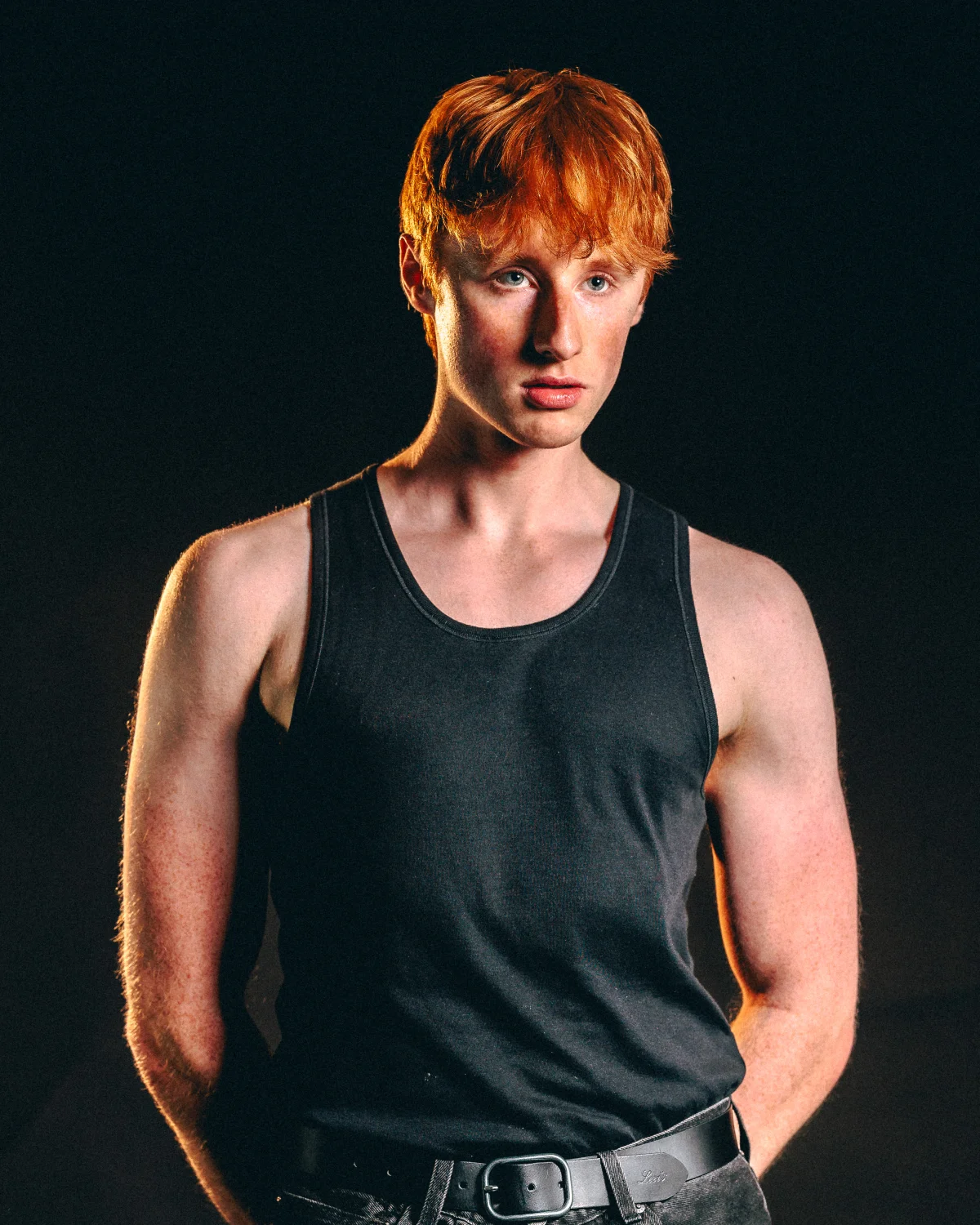 Young man with red hair in a black tank top posing against a dark background with dramatic lighting.