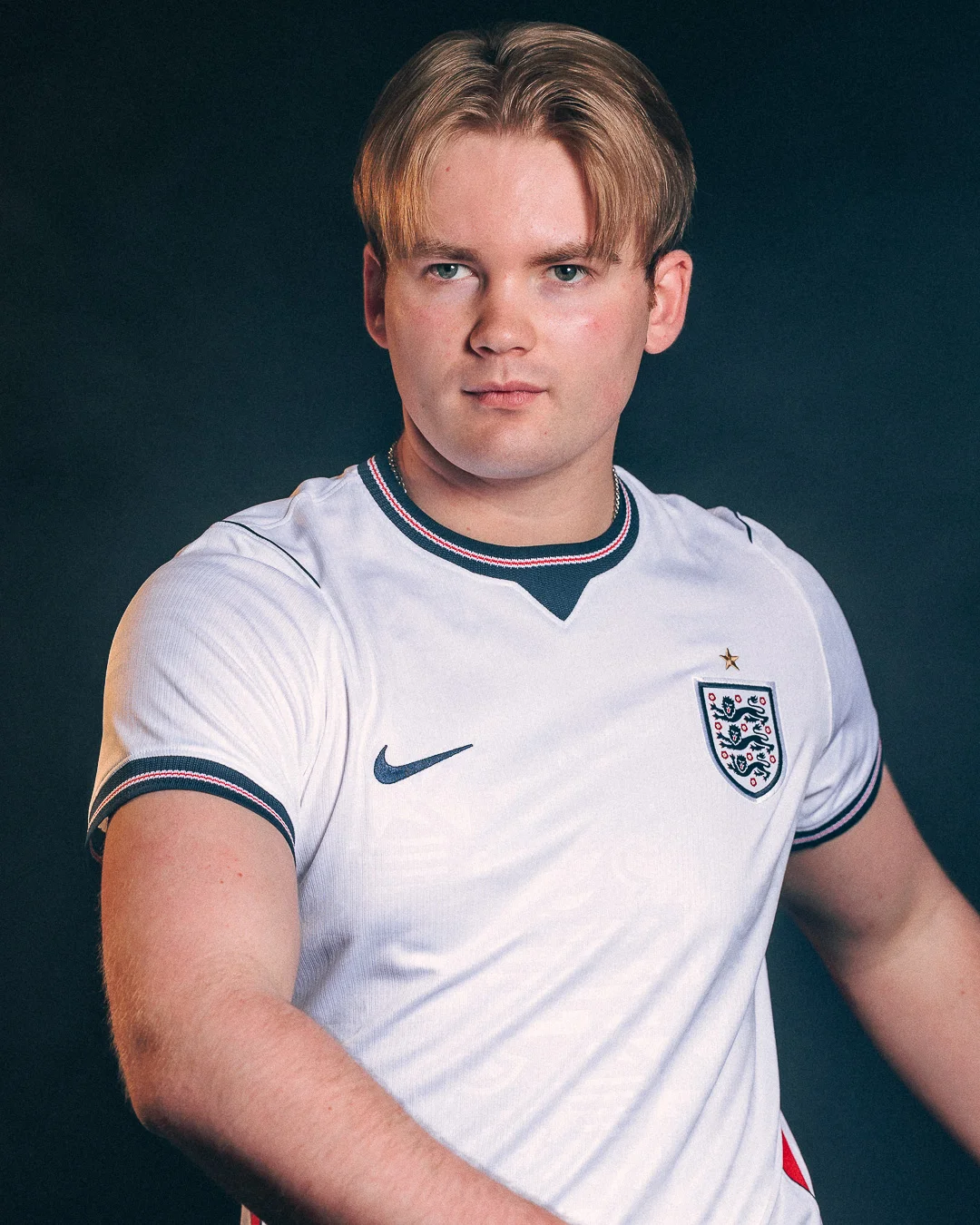 A studio portrait of a young man with blond hair wearing an England football jersey against a dark blue background.