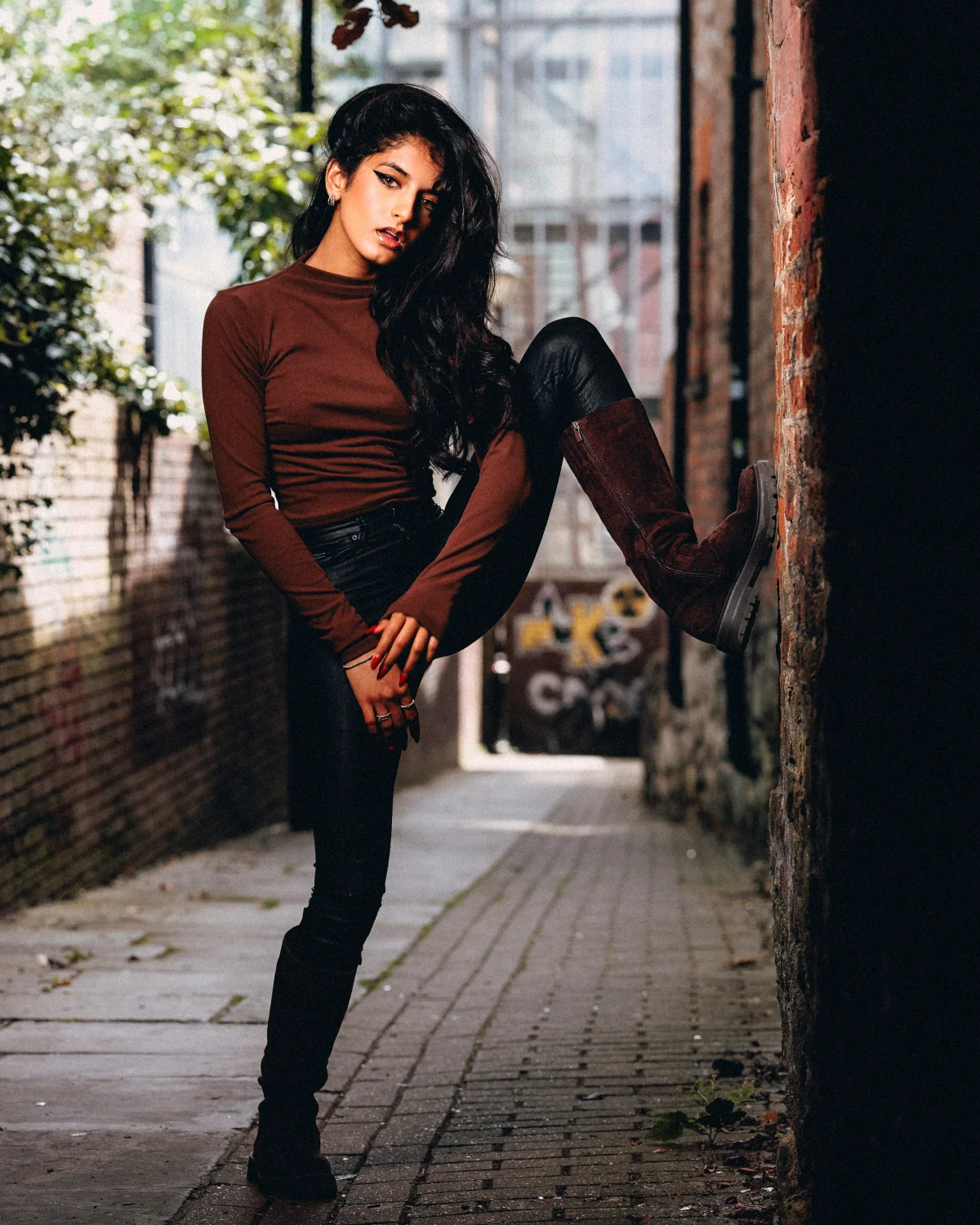 Woman posing in an urban alley wearing a brown top, black leather pants, and tall brown boots.